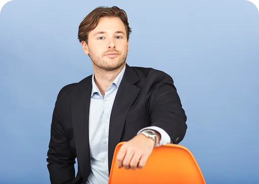 Portrait of a man with short brown hair and light stubble, wearing a dark blazer and light blue shirt. He sits against a plain blue background, resting one arm on the back of an orange chair and looking toward the camera.